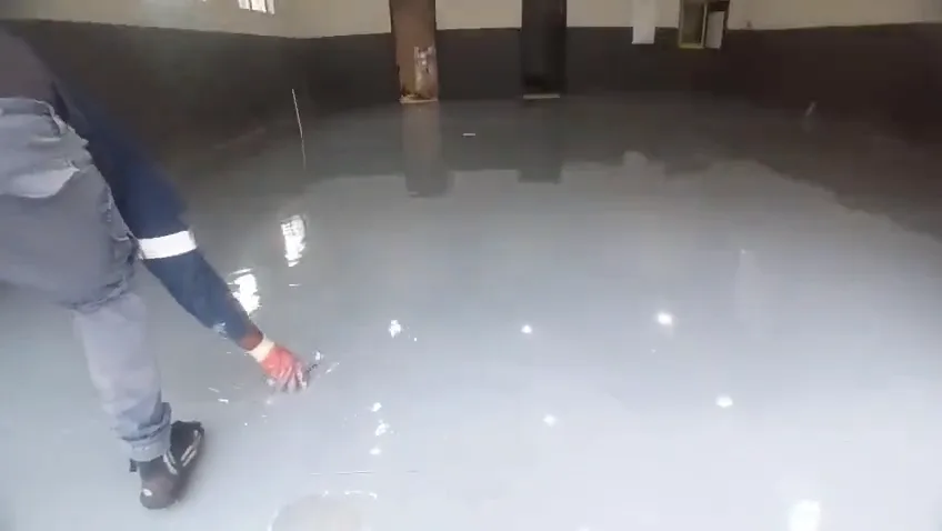 Installer in gloves spreading wet grey self-levelling epoxy across a commercial unit floor with a flat trowel
