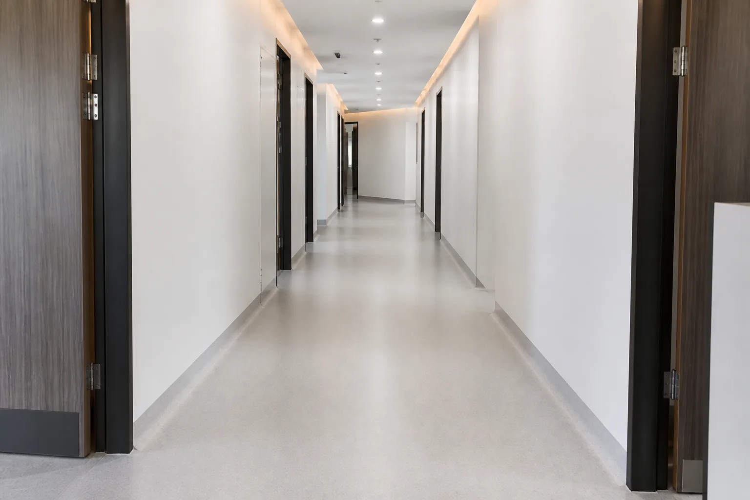 Long medical-suite corridor with a seamless light-grey speckled epoxy floor that curves up into coved skirting along white walls and dark doorframes