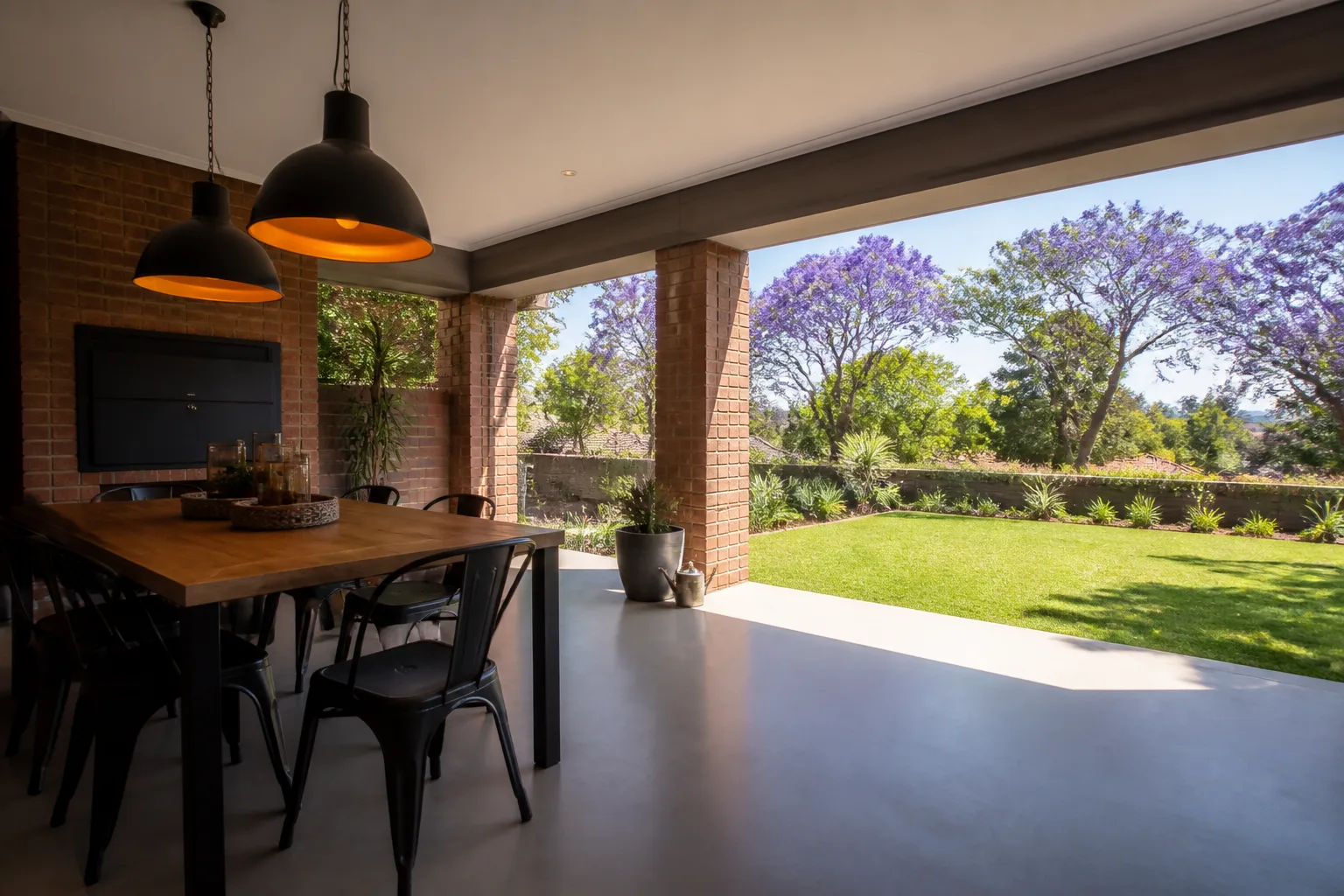 Covered patio with a smooth light-grey seamless floor, a wooden dining table with black metal chairs under pendant lights, a face-brick wall and a lawn with jacaranda trees in bloom